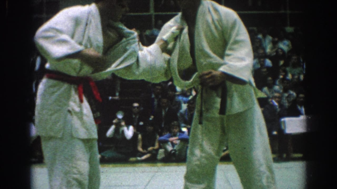 Two karate competitors in white gi grip each other as spectators watch behind them.