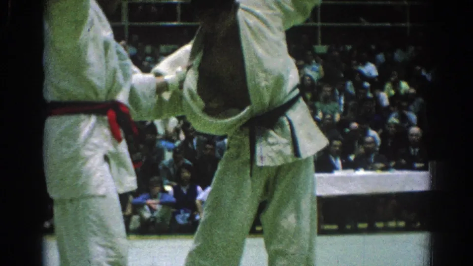 Karate competitors remain locked in a close technique with a seated crowd visible behind them in the 1966 San Gabriel footage.