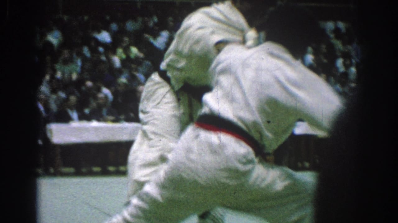 Two men in white gi grapple at close range during a 1966 judo match in San Gabriel, with a seated crowd behind them.