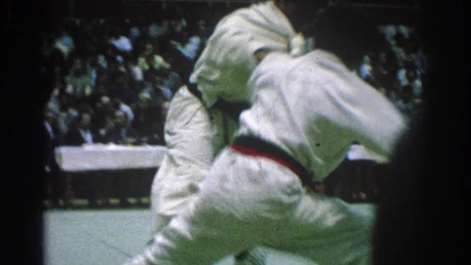 Two men in white gi grapple at close range during a 1966 judo match in San Gabriel, with a seated crowd behind them.