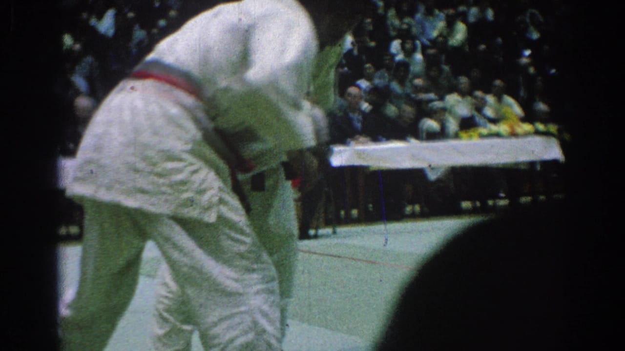 A judo competitor bends forward as his opponent presses in during the San Gabriel tournament bout, with spectators in the background.