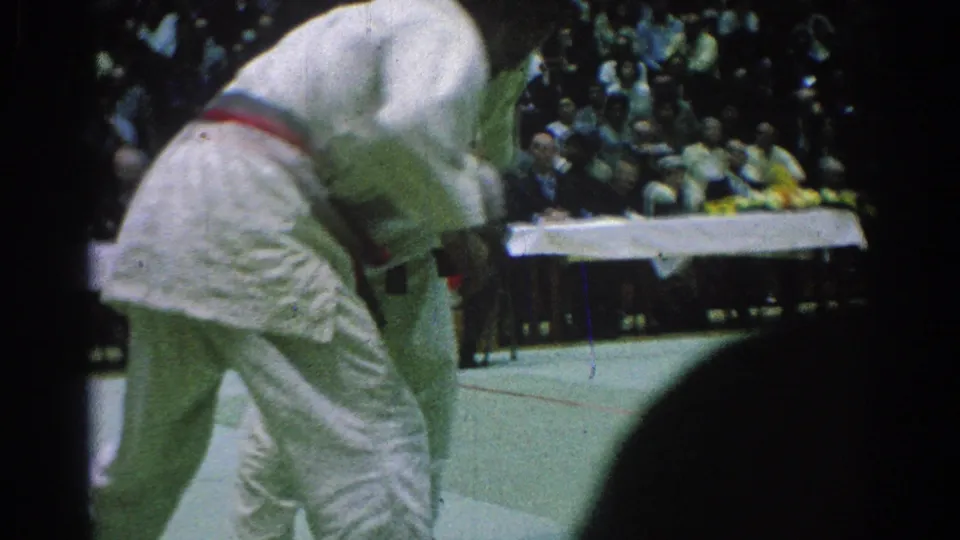 A judo competitor bends forward as his opponent presses in during the San Gabriel tournament bout, with spectators in the background.