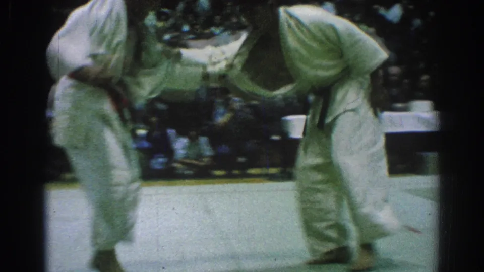 The two martial artists stay locked in a low stance during the 1966 judo match, filmed from a handheld spectator angle.