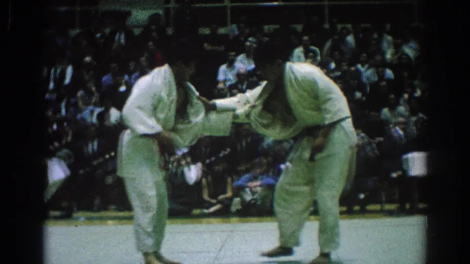 Two competitors in white gi face each other and appear to bow or reset during the San Gabriel judo tournament, with spectators behind them.