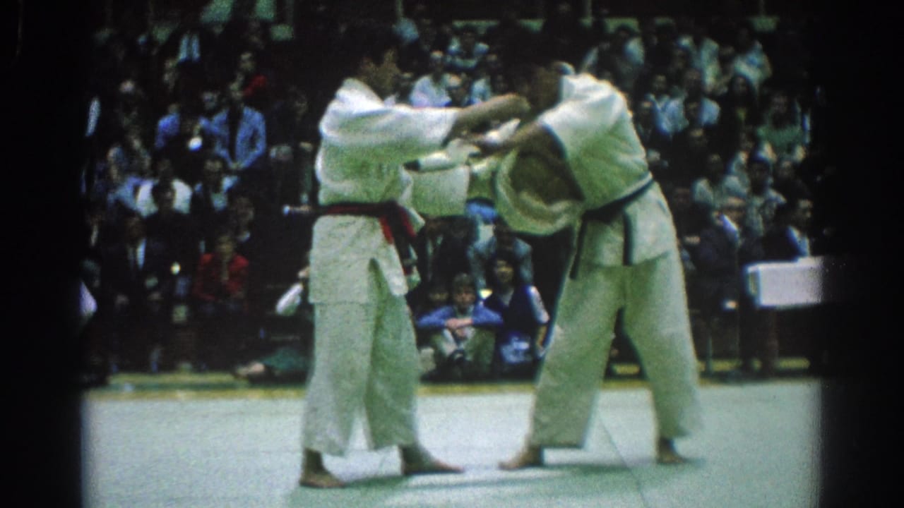 Two judo competitors in white gis grip each other on a mat before seated spectators.