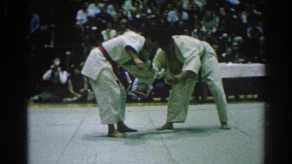 The two competitors remain locked in grips on the tatami, bending forward under bright indoor light with spectators behind them.
