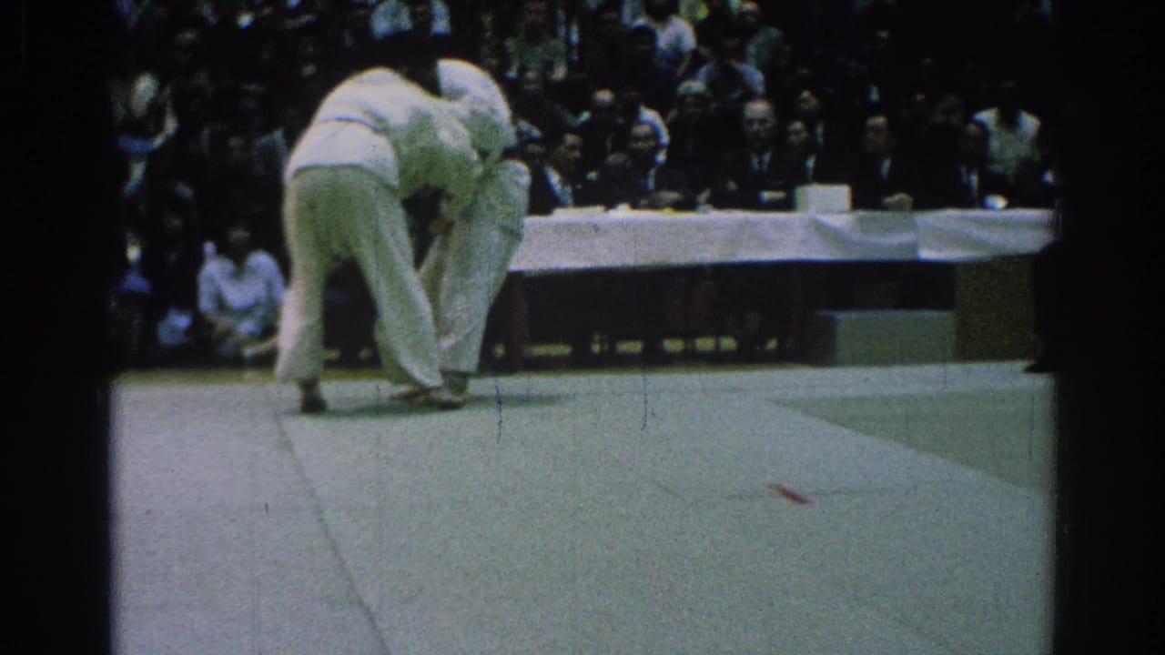 Two judokas in white gi grapple on a mat before seated judges and spectators.
