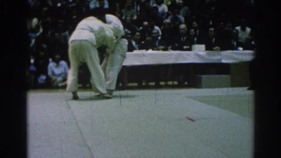 Two judokas compete on a mat during a 1966 San Gabriel judo match, with judges at a table and spectators behind them.