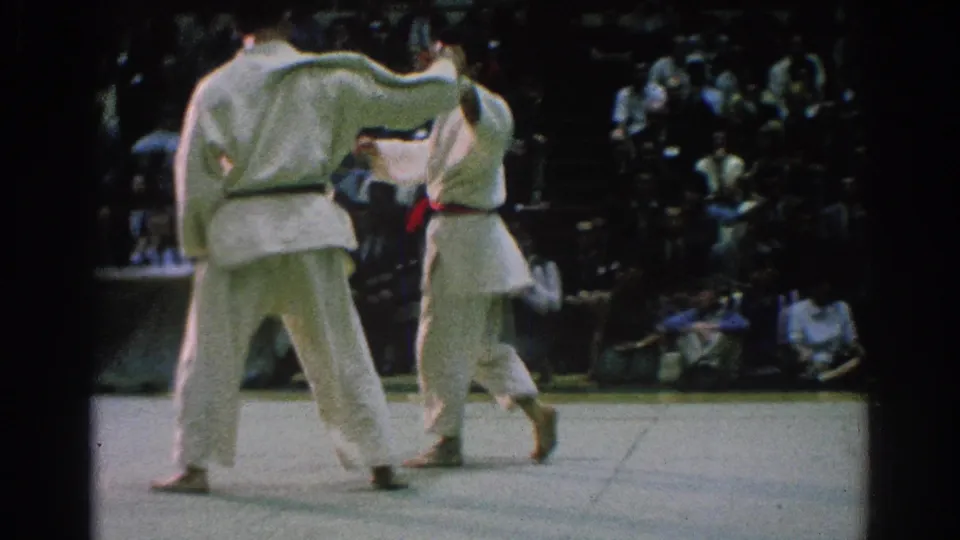 Two competitors reset or close distance during the 1966 judo bout, framed from behind with a seated audience in the background.