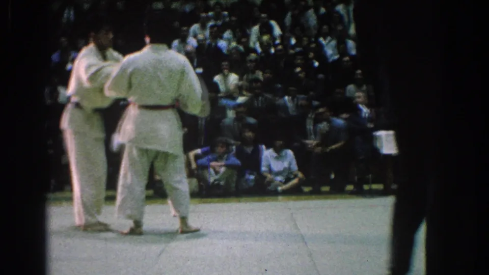The judo match pauses or resets in San Gabriel, with competitors on the mat, spectators behind them, and a figure partly visible at right.