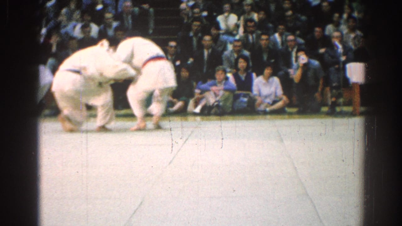 Two judo athletes grapple on a mat before a seated crowd.