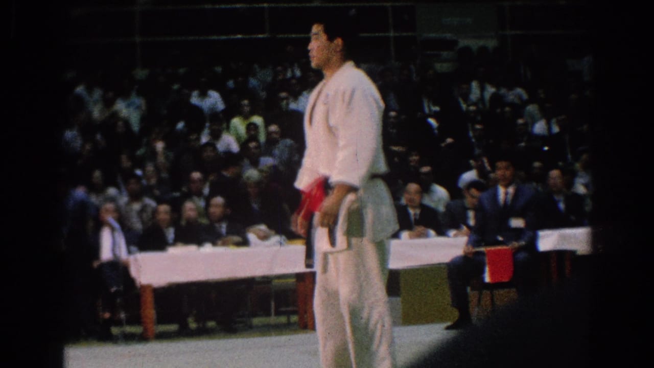 A red-belt judo athlete stands on the indoor competition mat with a crowd and officials behind him in San Gabriel footage.