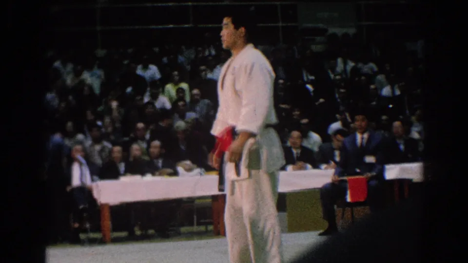 A red-belt judo athlete stands on the indoor competition mat with a crowd and officials behind him in San Gabriel footage.