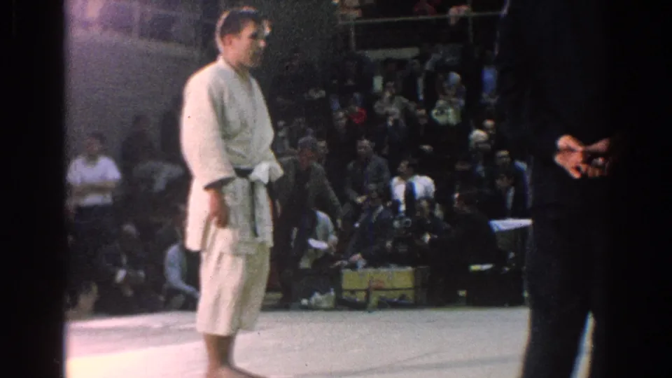 A judo competitor in a white gi stands near the mat edge as the audience fills the background in this 1960s gymnasium scene.