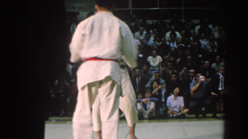 A judo athlete with a red belt stands with his back to the camera while another competitor and spectators are visible on the mat.
