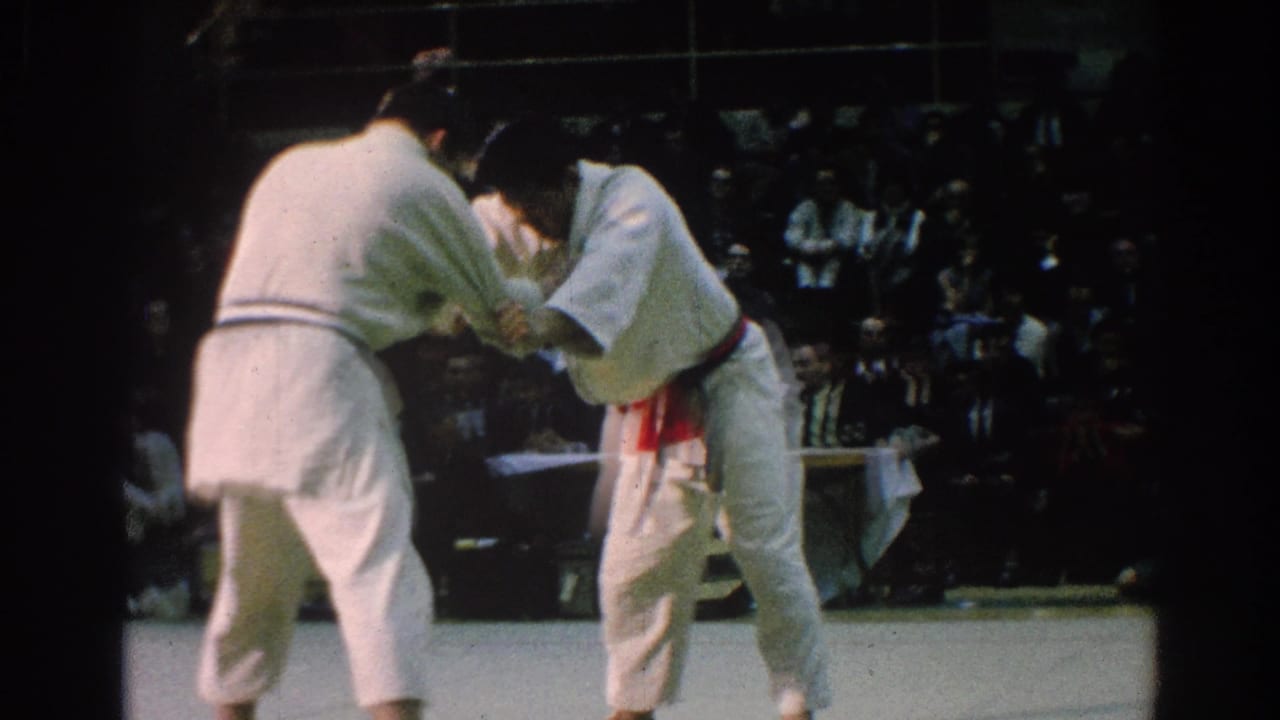 Two martial artists in white gi grip each other on a competition mat as spectators watch behind them.