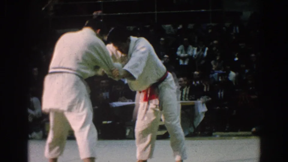 In San Gabriel, California, 1966, two karate competitors in white gi hold a close clinch on a pale mat before a seated crowd.