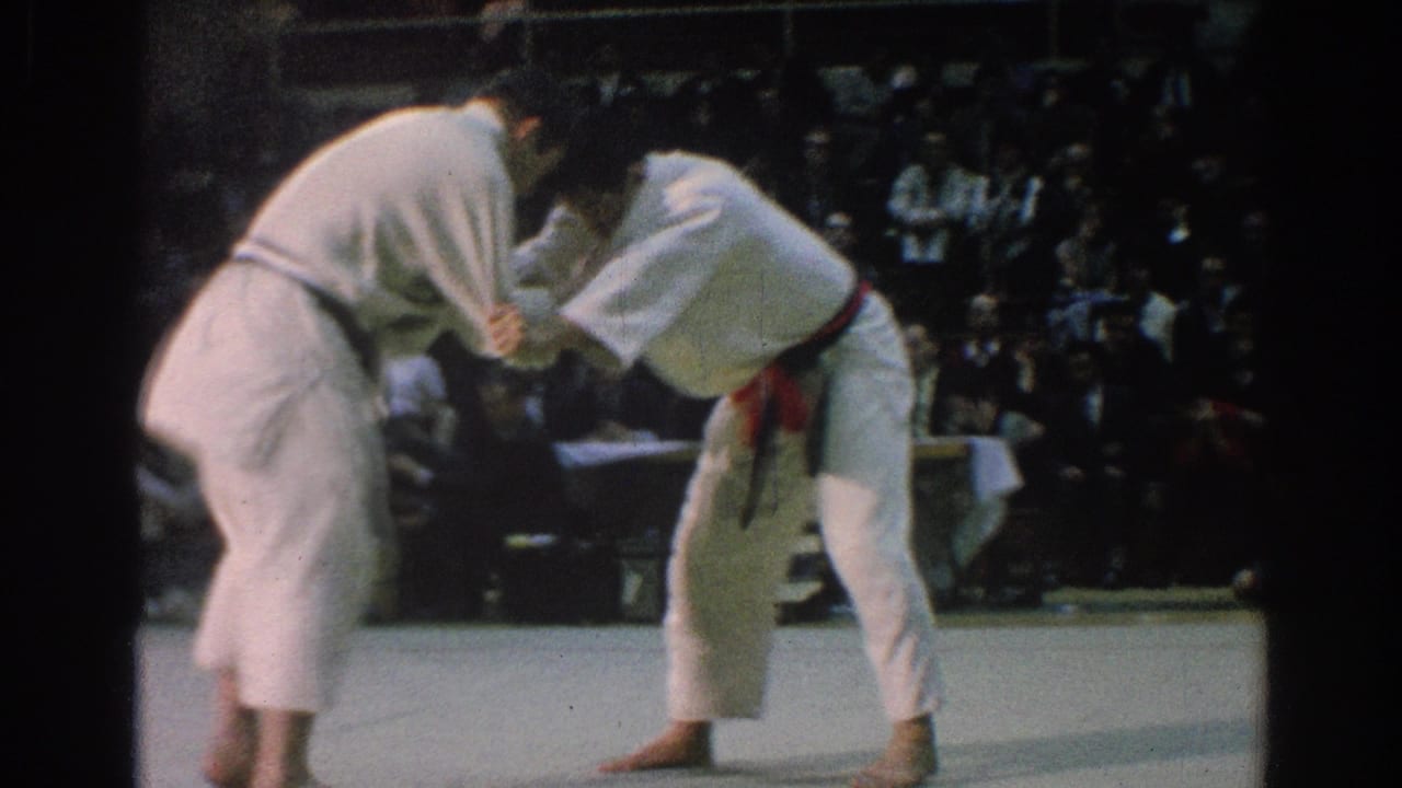 Two competitors in white uniforms remain locked together on a mat with spectators seated in the background.
