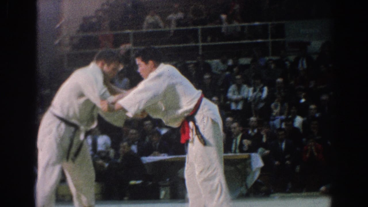 Two judokas in white gi grip each other on a mat before a seated crowd.