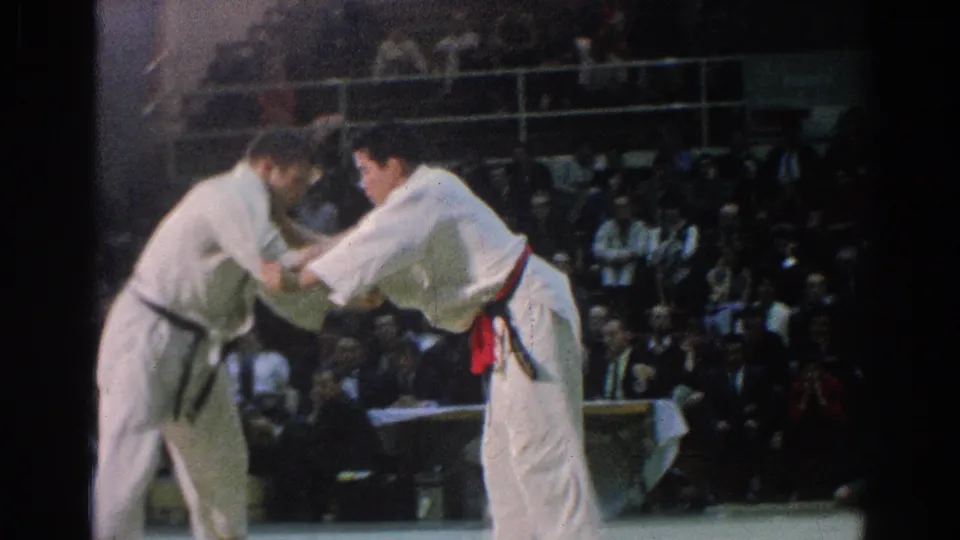 Two competitors in white gi begin a judo exchange in a 1966 San Gabriel indoor arena, with spectators visible behind them.