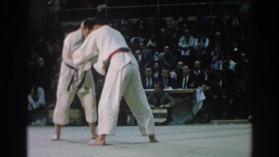 The two judokas remain locked in close grips during the 1966 indoor match, with the arena audience watching from tiered seating.