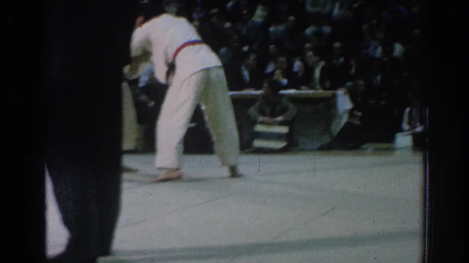 A 1966 San Gabriel karate bout continues as two gi-clad competitors move across the mat before judges and spectators.