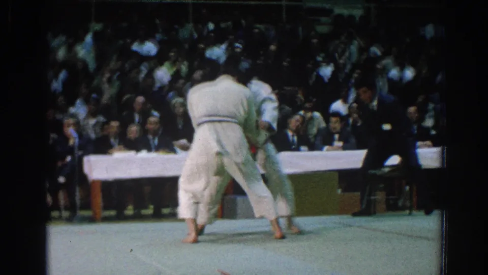 Two men in white gi grapple or close distance during a 1966 karate match, filmed from behind near the audience side.
