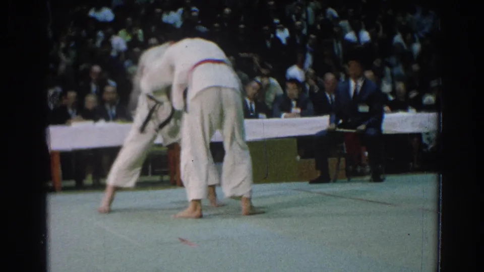 The 1966 San Gabriel karate match shows two competitors in white gi moving near the officials table before a packed audience.