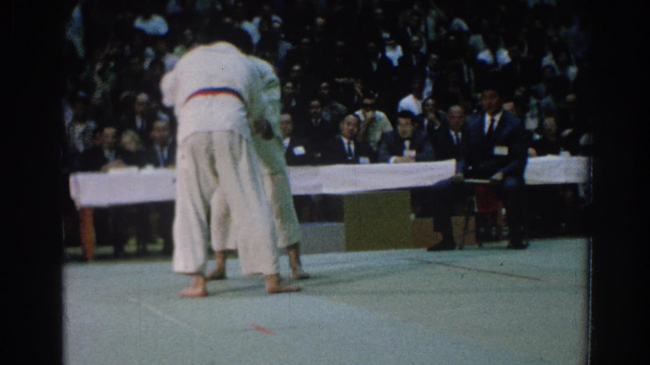 Two martial artists in white gi grapple on a mat before spectators.