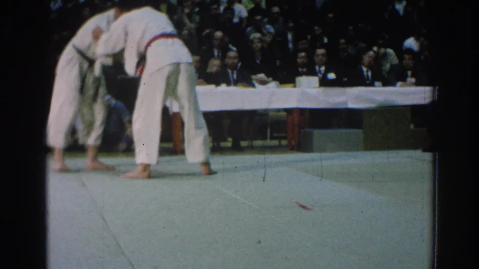 The martial artists remain locked in close contact on the mat at the 1966 San Gabriel tournament, with judges and spectators in the background.