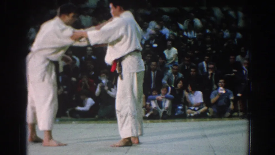Two men in white gi face each other near the end of the 1966 San Gabriel match clip, with a seated crowd visible behind them.