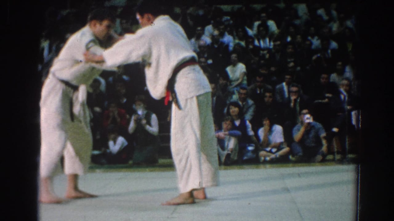 In 1966 San Gabriel, California footage, two men in white gi and colored belts begin grappling on a gymnasium mat before an audience.