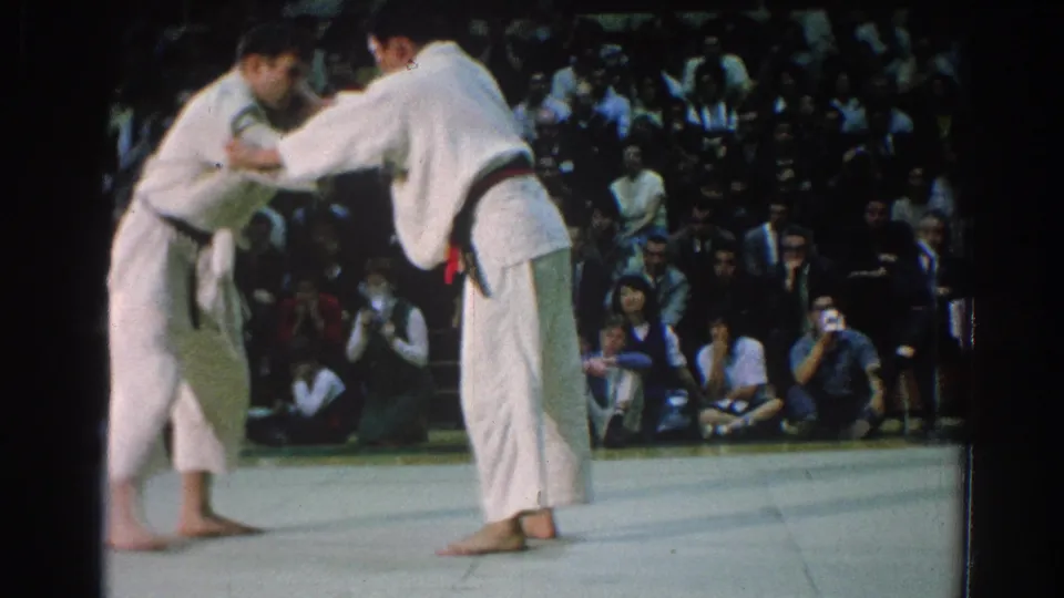 In 1966 San Gabriel, California footage, two men in white gi and colored belts begin grappling on a gymnasium mat before an audience.