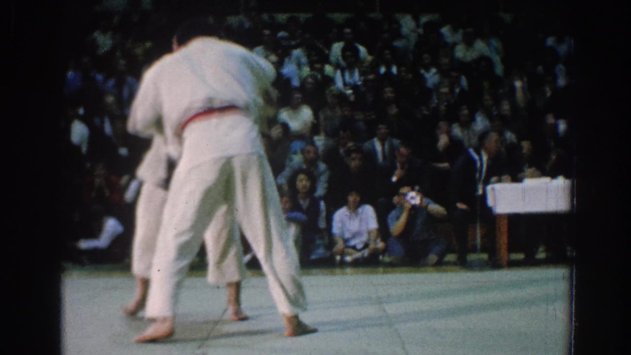 The match continues on a gymnasium mat, with one competitor turned away from the camera and spectators visible behind them.