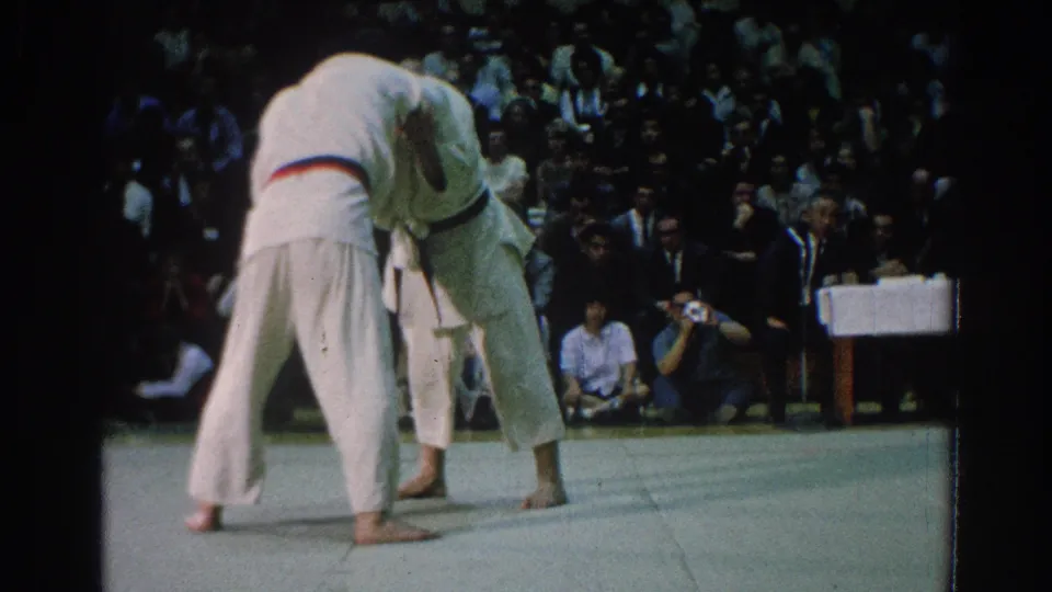 A gi-clad competitor leans into a throwing position while his opponent stays close, with the audience watching from behind.