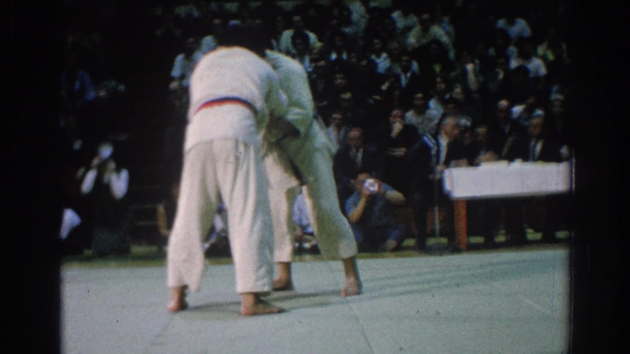 Two judo competitors in white uniforms grapple on a mat before seated spectators.