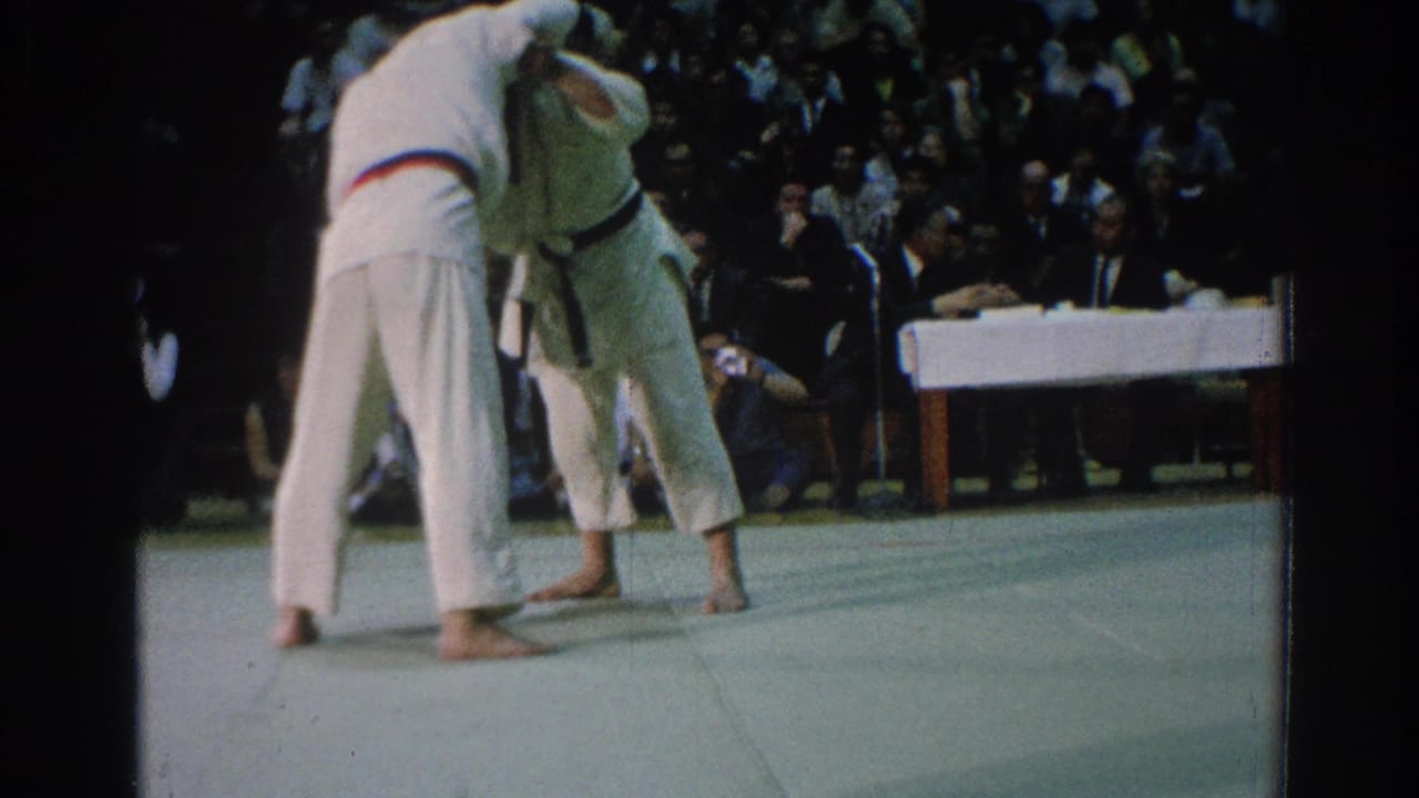 The judo competitors hold a standing clinch during the San Gabriel match, with the audience and officials visible beyond the mat.