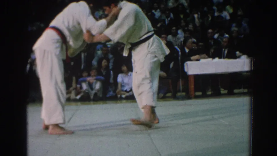 In the 1966 San Gabriel judo scene, the competitors lean forward in a close grip as spectators sit behind the mat.