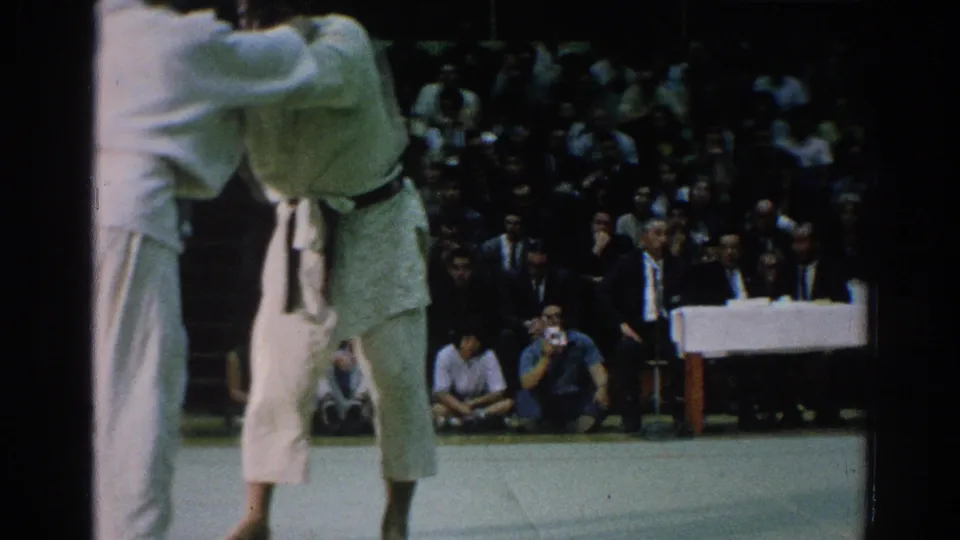 The handheld 8mm frame crops one competitor at left while the other stands in a black belt on the San Gabriel judo mat.