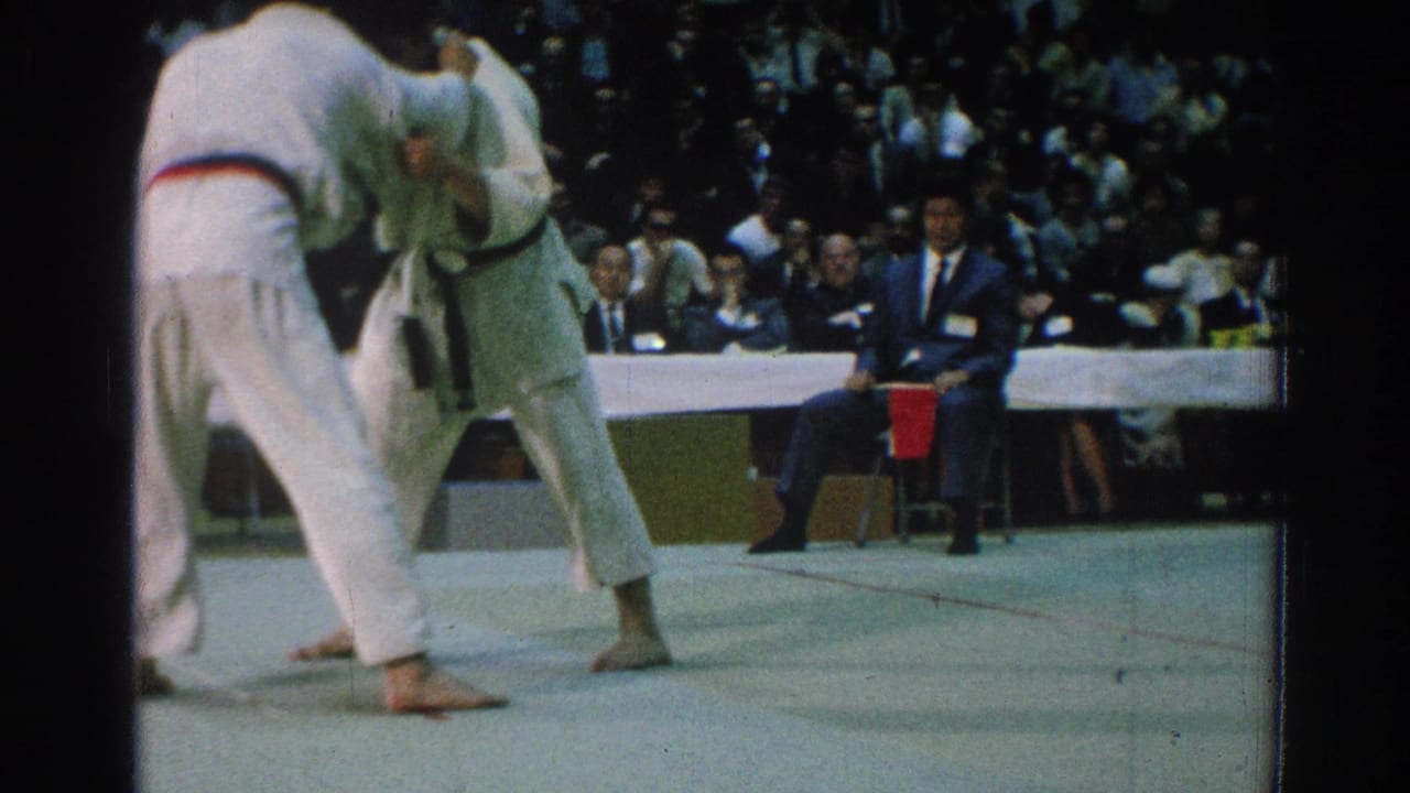 Two men in white gi grapple on a mat before seated officials and an audience.