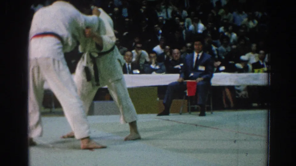 Two martial arts competitors in white gi grapple on a mat during a 1966 San Gabriel, California event as officials and spectators watch.