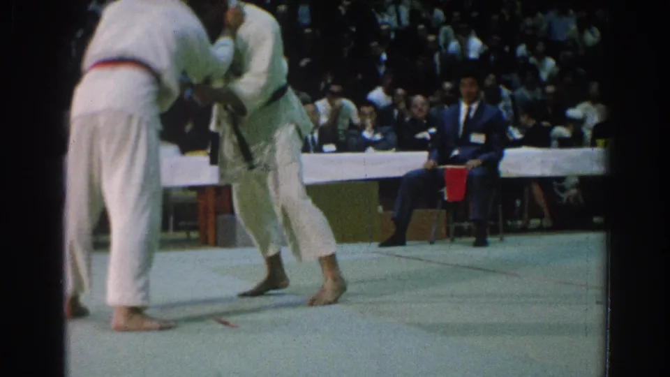 A 1966 San Gabriel martial arts match shows two men in white gi locked in a standing grapple before judges and spectators.