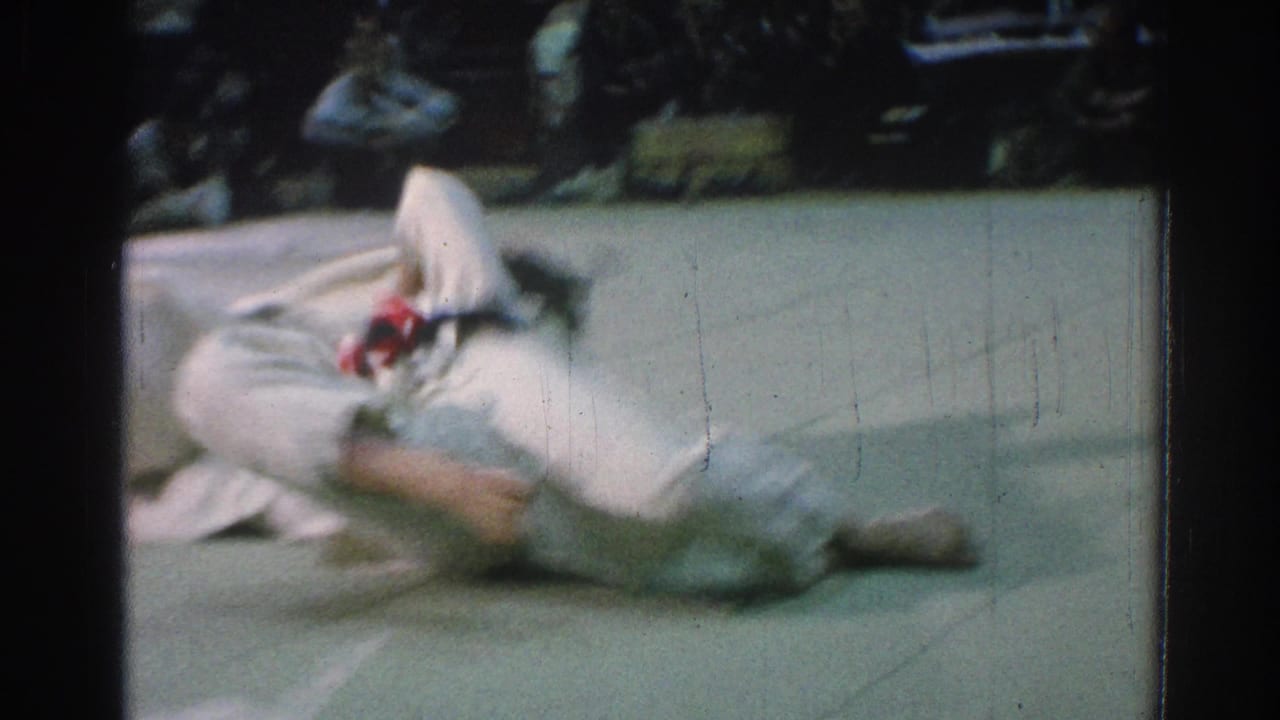 Two martial artists in white gi uniforms grapple on a mat as one falls.
