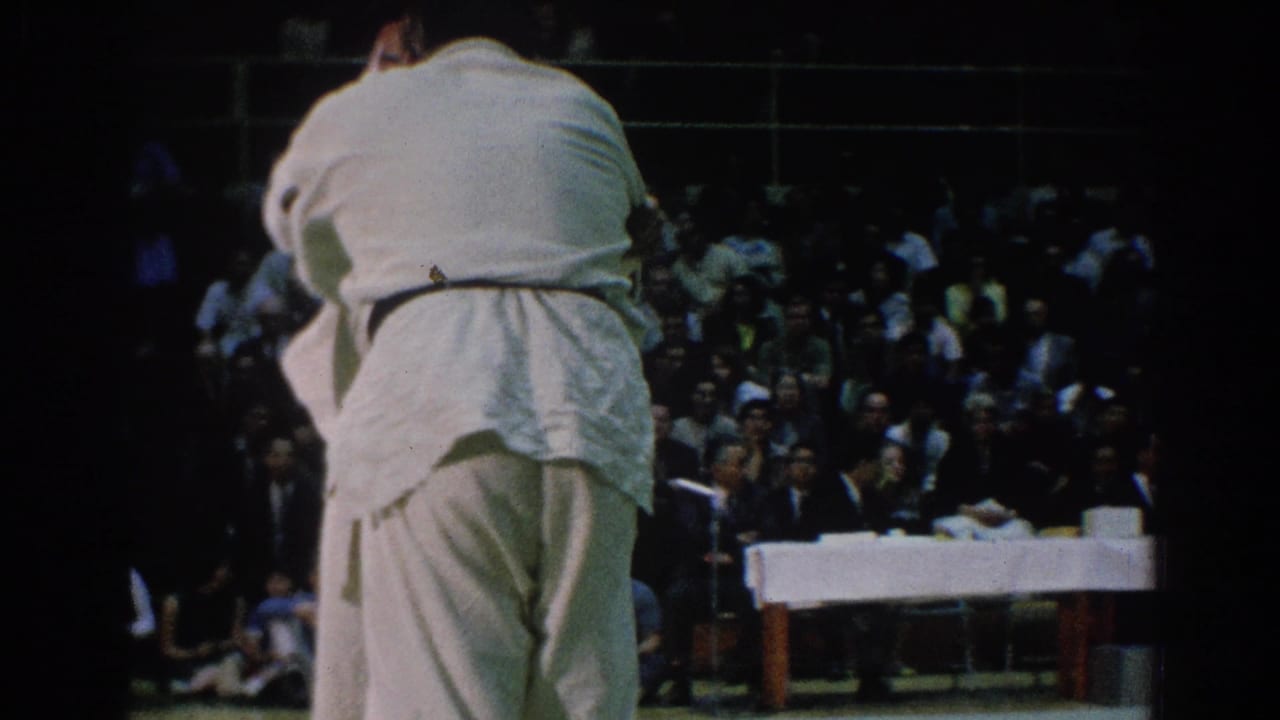 A martial artist in a white gi stands with back to camera before spectators.