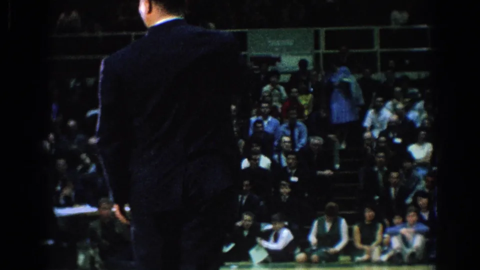 A suited official or judge stands beside the competition area as spectators watch the 1966 indoor martial arts match.