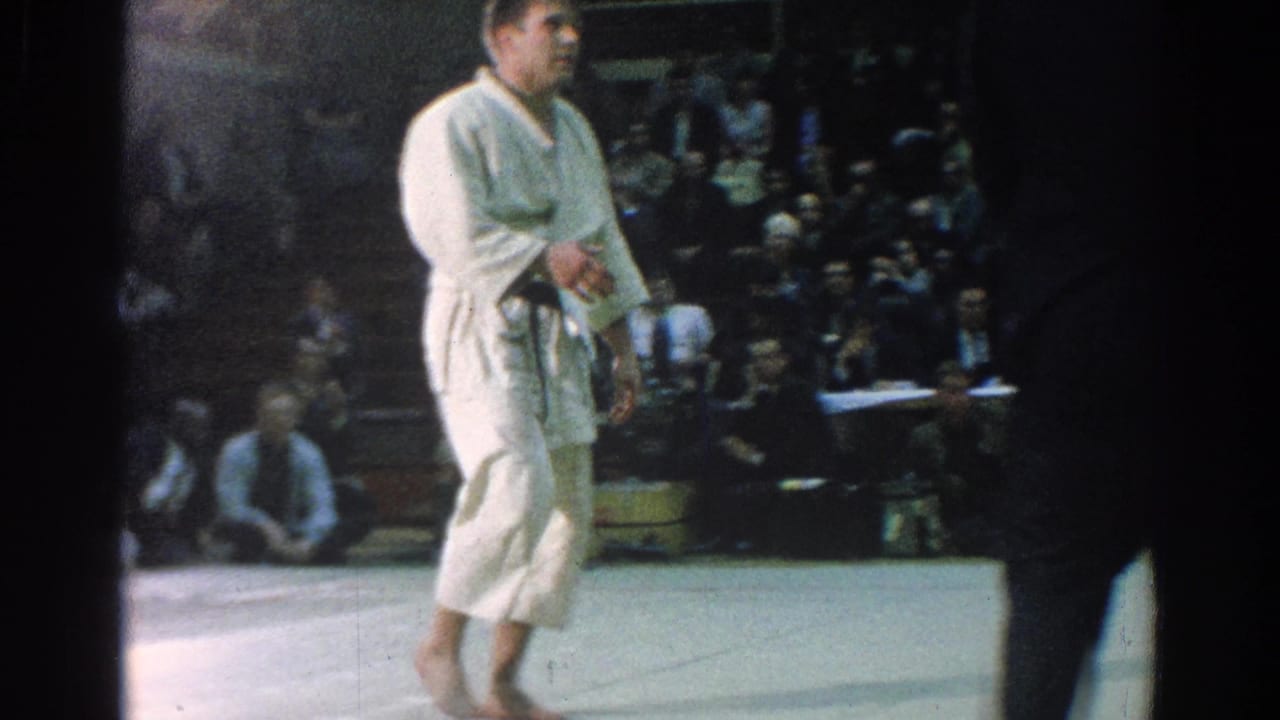 Karate competitor in a white gi stands on a mat before a seated crowd.
