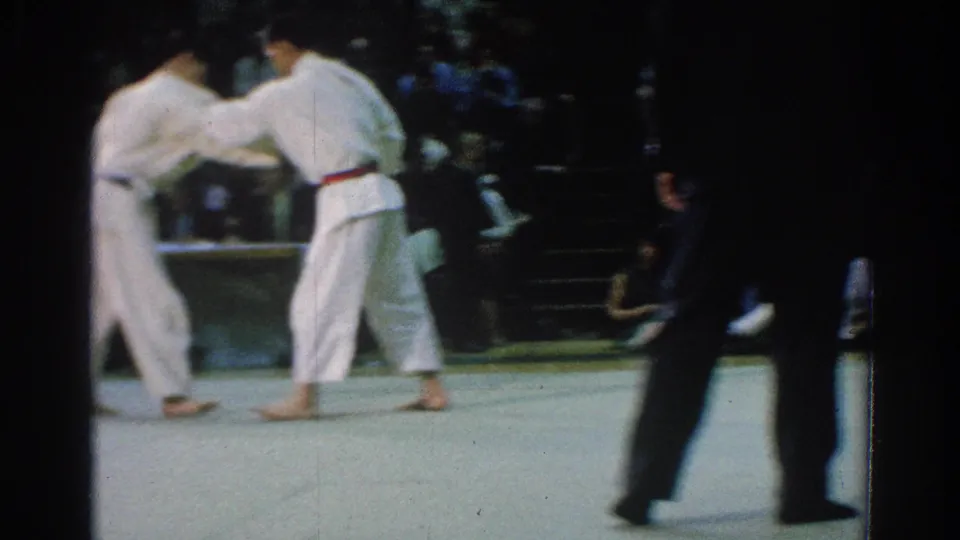 A handheld 8mm frame catches two white-gi competitors grappling while the referee passes in the foreground.