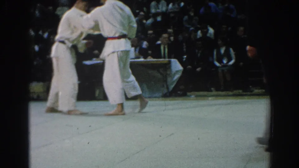 Two competitors in white gi remain in close contact on the tournament mat, watched by a crowded indoor audience.