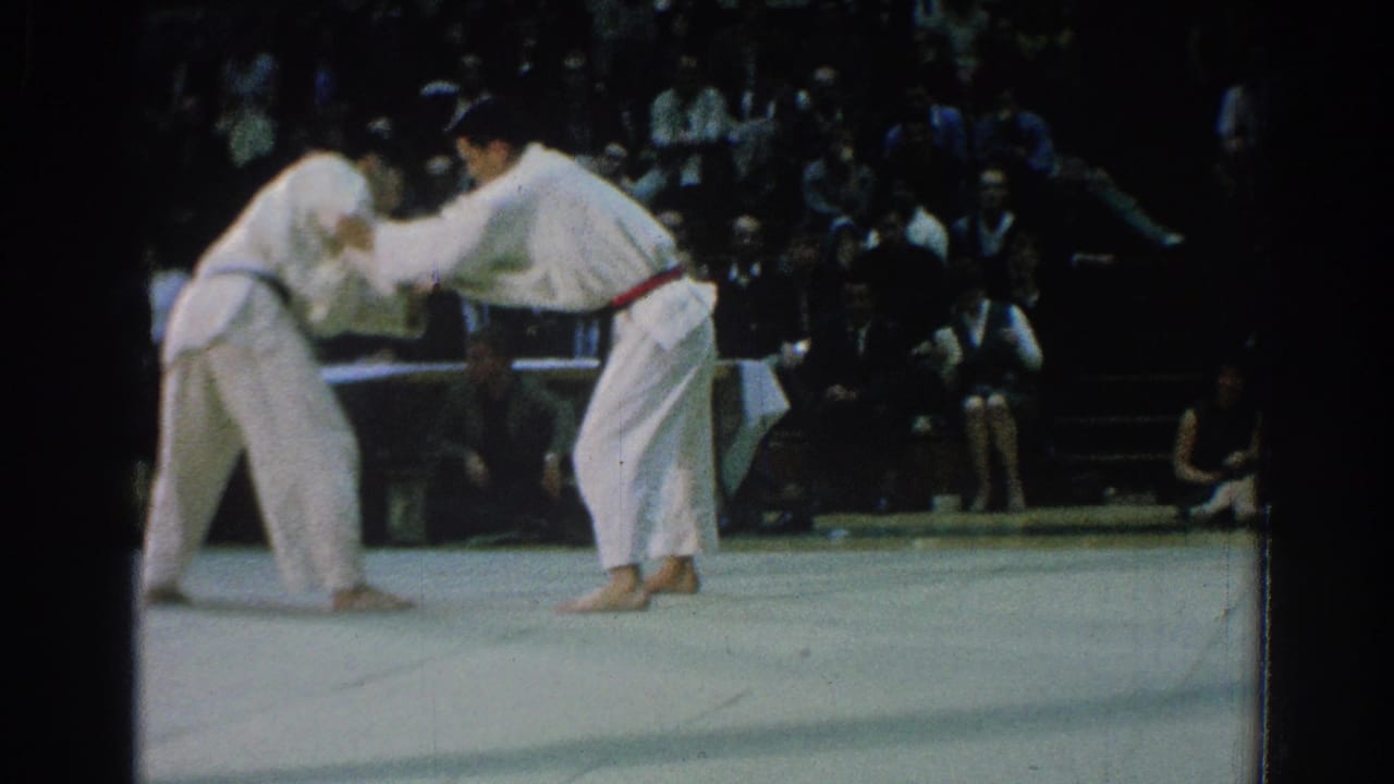 Two karate competitors in white gi grip each other on a mat before a seated crowd.