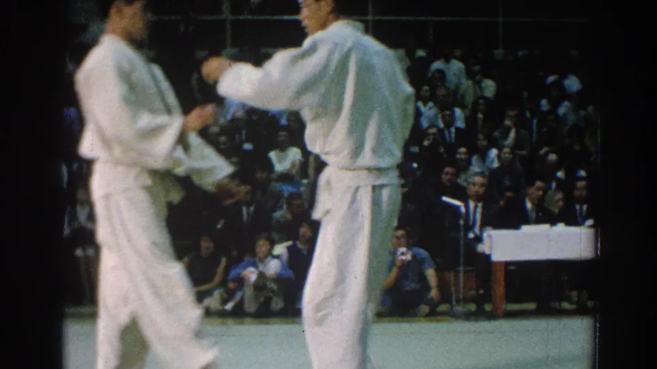 Two male karate competitors reset or circle on the mat during the 1966 San Gabriel indoor tournament footage.